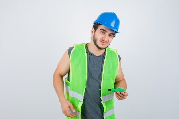 Homme jeune constructeur tenant un tournevis tout en regardant ailleurs en uniforme et à la vue de face, confiant.