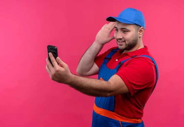 Homme jeune constructeur en uniforme de construction et cap holding smartphone lookingat écran connexion avec quelqu'un agitant avec salutation de la main