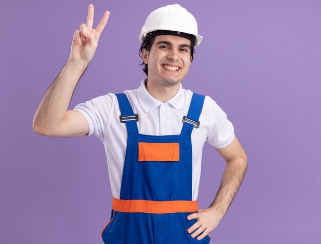 Homme jeune constructeur en uniforme de construction et casque de sécurité à l'avant souriant avec visage heureux montrant v-sign debout sur mur violet