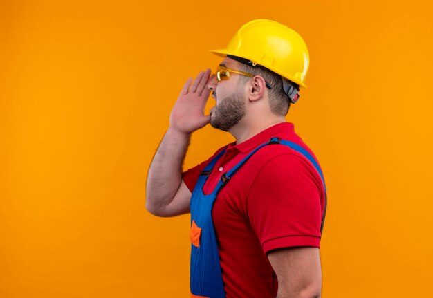 Homme jeune constructeur en uniforme de construction et casque de sécurité sur le côté en criant ou en appelant quelqu'un avec la main près de la bouche