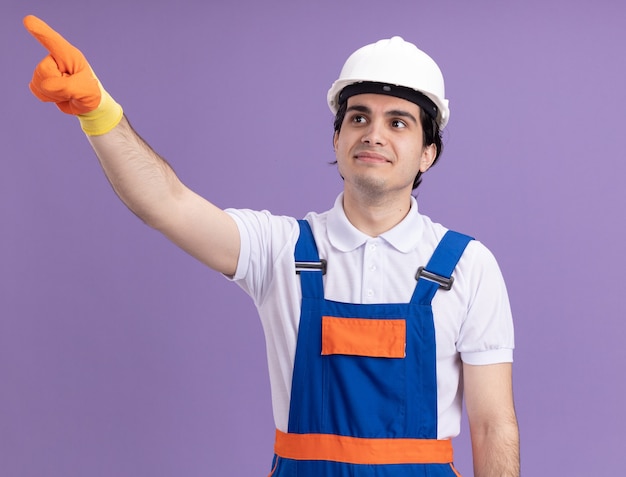 Homme jeune constructeur en uniforme de construction et casque de sécurité dans des gants en caoutchouc pointant avec l'index à quelque chose de souriant debout sur le mur violet