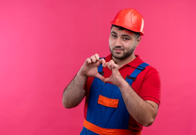 Homme jeune constructeur en uniforme de construction et casque de sécurité faisant le geste du cœur avec les doigts sur la poitrine en souriant