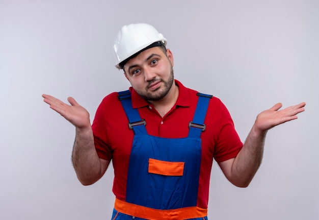 Homme jeune constructeur en uniforme de construction et casque de sécurité à haussant les épaules confus et incertain n'ayant pas de réponse