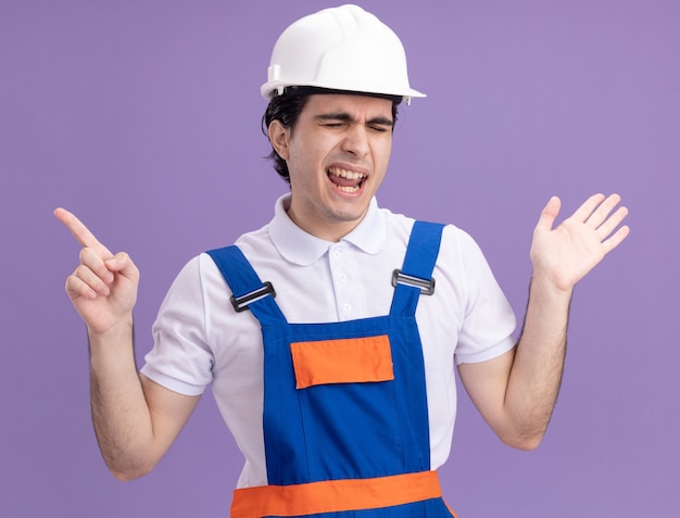 Homme jeune constructeur en uniforme de construction et casque de sécurité heureux et joyeux pointant avec l'index sur le côté debout sur le mur violet