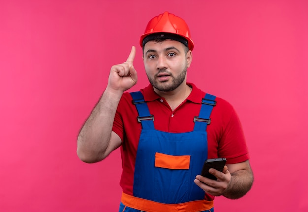Homme jeune constructeur en uniforme de construction et casque de sécurité holding smartphone montrant l'index en se rappelant de ne pas oublier