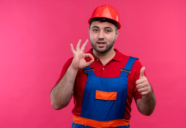 Homme jeune constructeur en uniforme de construction et casque de sécurité montrant les pouces vers le haut faisant signe ok