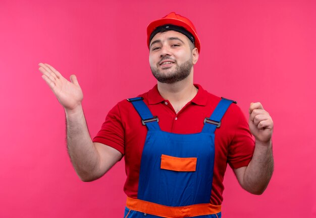 Homme jeune constructeur en uniforme de construction et casque de sécurité pointant avec le bras de sa main sur le côté à la recherche de sourire confiant