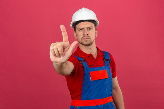 Homme jeune constructeur en uniforme de construction et casque de sécurité pointant avec le doigt vers le haut et expression en colère sur mur rose isolé