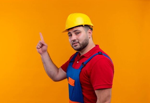 Homme jeune constructeur en uniforme de construction et casque de sécurité pointant avec l'index jusqu'à la recherche intelligente et confiante axée sur la tâche