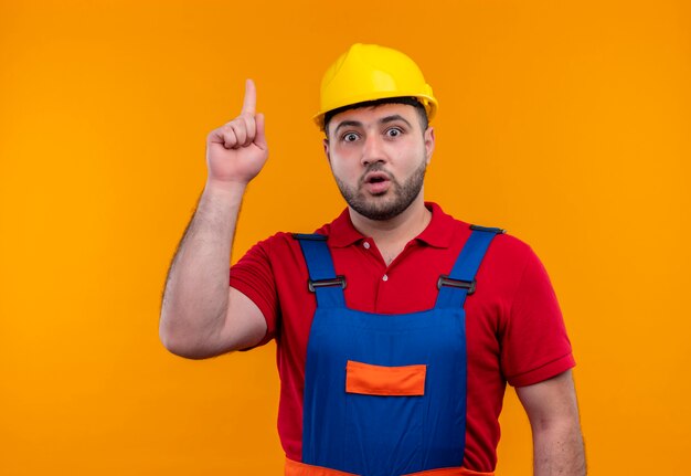 Homme jeune constructeur en uniforme de construction et casque de sécurité pointant avec l'index à la surprise