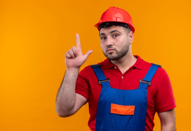 Homme jeune constructeur en uniforme de construction et casque de sécurité à la surprise montrant l'index