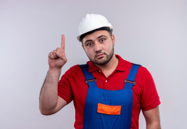 Homme jeune constructeur en uniforme de construction et casque de sécurité à la surprise de pointer avec l'index