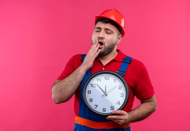 Homme jeune constructeur en uniforme de construction et casque de sécurité tenant horloge murale bâillement à la fatigue