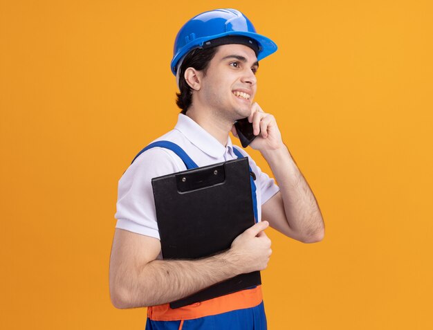 Homme jeune constructeur en uniforme de construction et casque de sécurité tenant le presse-papiers souriant parler sur téléphone mobile debout sur un mur orange