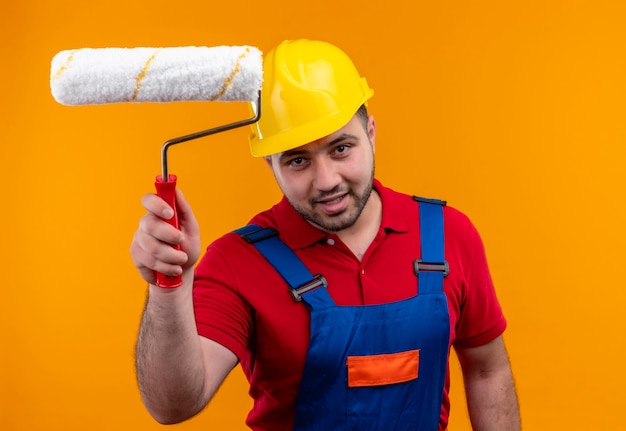 Photo gratuite homme jeune constructeur en uniforme de construction et casque de sécurité tenant un rouleau à peinture souriant confiant