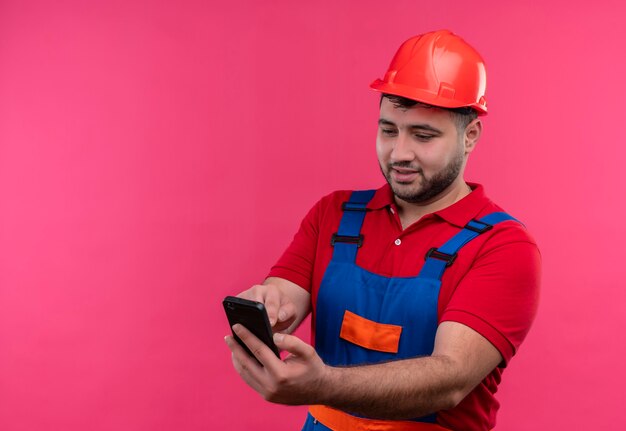 Homme jeune constructeur en uniforme de construction et casque de sécurité tenant un téléphone mobile à la recherche de plaisir et positif