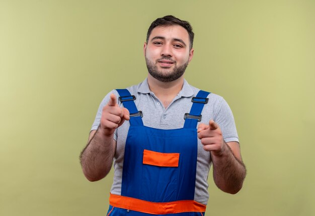 Homme jeune constructeur en uniforme de construction pointant avec l'index à la caméra souriant confiant