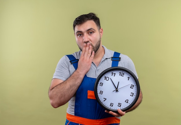 Photo gratuite homme jeune constructeur en uniforme de construction tenant horloge murale choqué couvrant la bouche avec la main