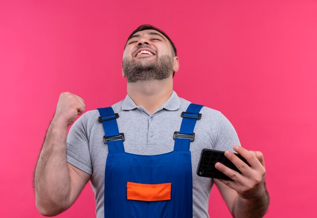 Homme jeune constructeur en uniforme de construction tenant le poing serrant le smartphone heureux et sorti