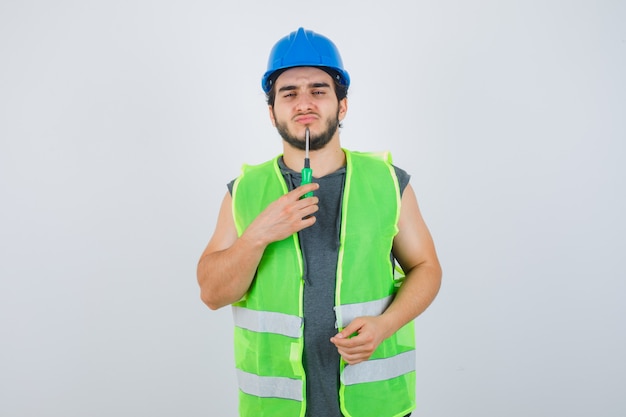 Homme jeune constructeur en uniforme tenant la pointe du tournevis sur le menton et à la vue réfléchie, de face.