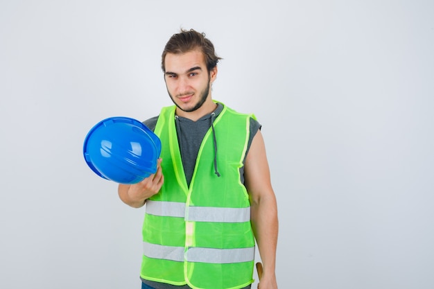 Homme jeune constructeur en uniforme de vêtements de travail tenant un casque et à la vue de face, confiant.