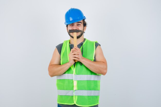 Homme jeune constructeur en uniforme de vêtements de travail tenant un marteau sur la bouche et à la drôle, vue de face.