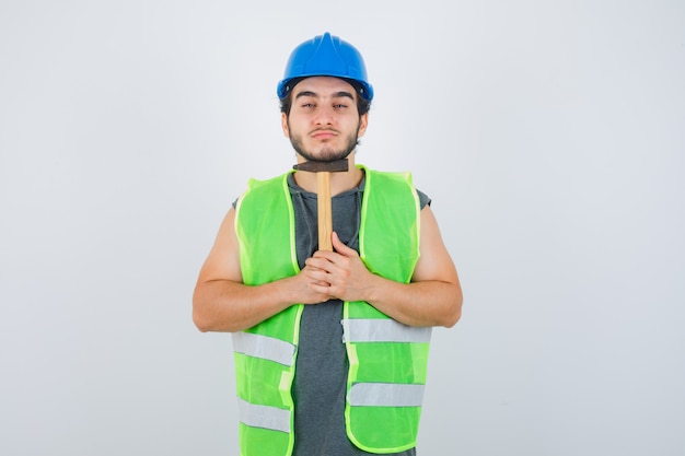 Homme jeune constructeur en uniforme de vêtements de travail tenant un marteau sous le menton et à la vue de face, confiant.