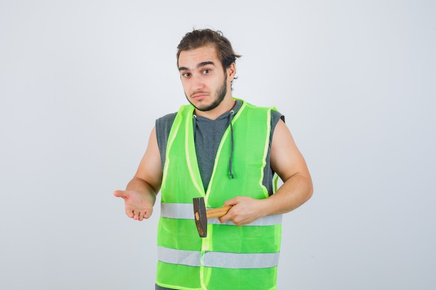 Homme jeune constructeur en uniforme de vêtements de travail tenant un marteau tout en montrant quelque chose et à la vue indécise, de face.