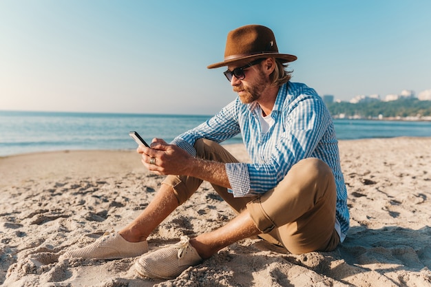 Homme jeune hipster attrayant assis sur la plage au bord de la mer en vacances d'été