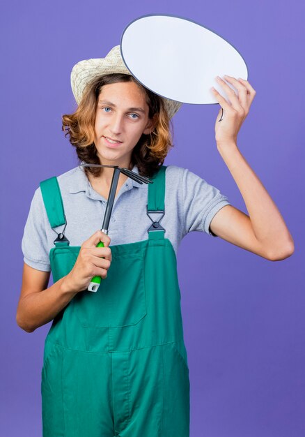 Homme jeune jardinier portant combinaison et chapeau tenant Mattock et bulle de dialogue vide