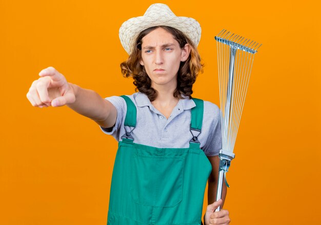 Homme jeune jardinier portant combinaison et chapeau tenant râteau pointant avec l'index