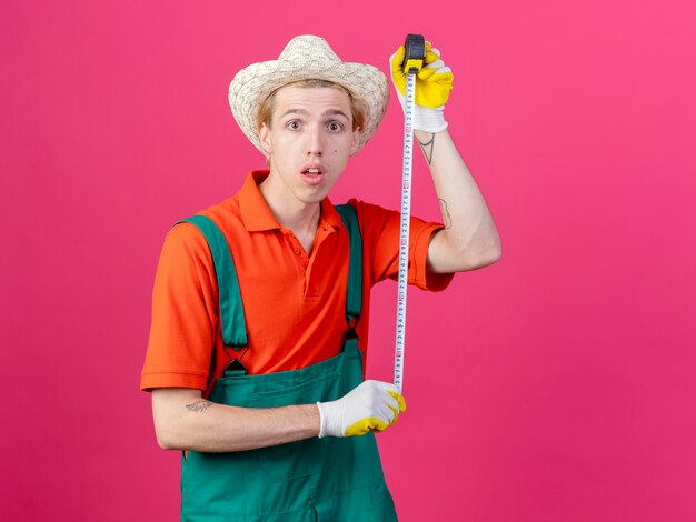 Homme jeune jardinier portant combinaison et chapeau tenant un ruban à mesurer inquiet