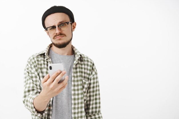 Homme jeune snob indifférent dans des lunettes et bonnet avec barbe tenant un smartphone souriant et regardant l'offre de réception insouciante et non impressionnée via des messages et être insatisfait