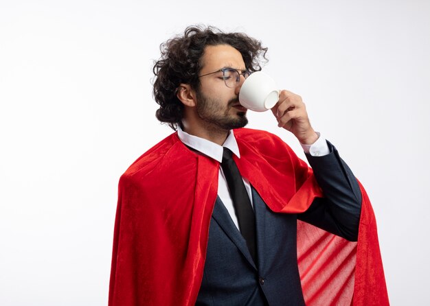 Homme jeune super-héros insatisfait dans des lunettes optiques portant costume avec cape rouge boissons de tasse isolé sur mur blanc