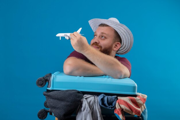 Homme jeune voyageur barbu en chapeau d'été avec valise pleine de vêtements tenant un avion jouet ludique optimiste et heureux souriant à côté avec un look de rêve 2