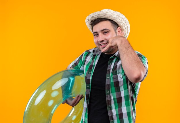 Homme jeune voyageur en chemise à carreaux et chapeau d'été tenant l'anneau gonflable souriant joyeusement faisant appelez-moi geste debout sur le mur orange