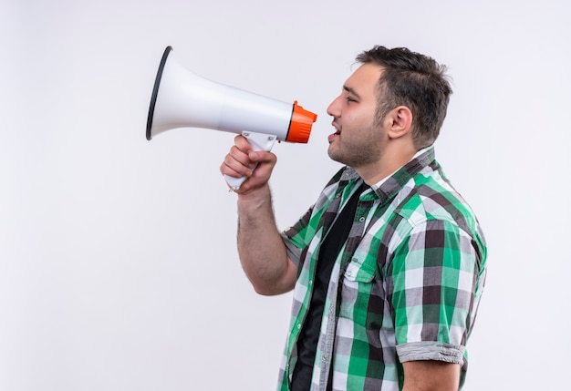 Homme jeune voyageur en chemise à carreaux criant au mégaphone debout sur un mur blanc