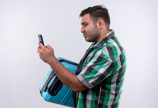 Homme jeune voyageur en chemise à carreaux debout avec valise regardant l'écran de son smartphone à la confusion sur mur blanc