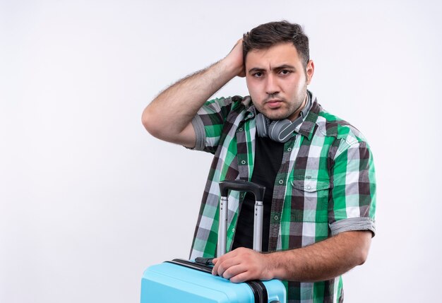 Homme jeune voyageur en chemise à carreaux holding valise à la tête de grattage incertain et confus debout sur un mur blanc