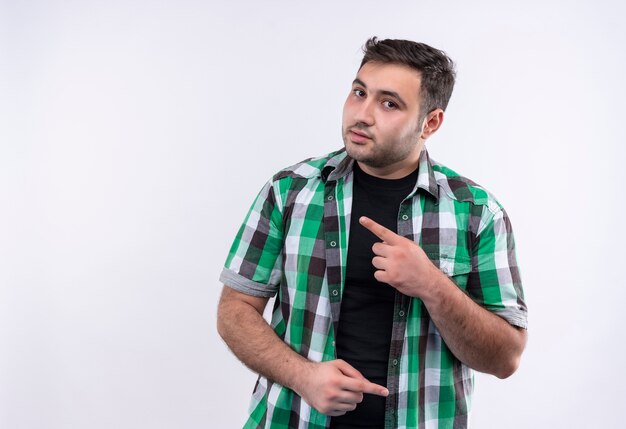 Homme jeune voyageur en chemise à carreaux pointant avec le doigt sur le côté avec une expression sérieuse confiante debout sur un mur blanc