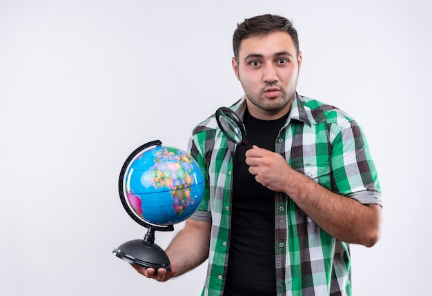 Homme jeune voyageur en chemise à carreaux tenant globe et loupe à la surprise debout sur un mur blanc