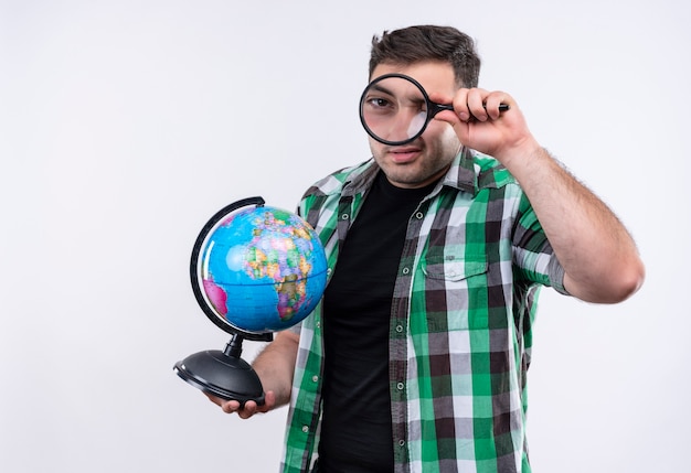 Homme jeune voyageur en chemise à carreaux tenant le globe à travers une loupe debout sur un mur blanc