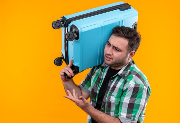 Homme jeune voyageur en chemise à carreaux tenant valise à côté, mécontent de se tenir debout sur un mur orange