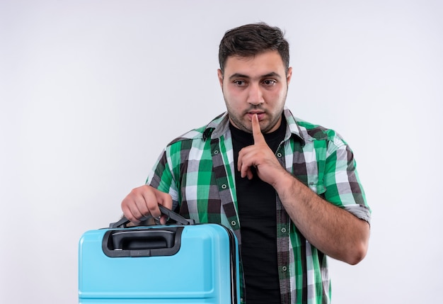 Homme jeune voyageur en chemise à carreaux tenant valise faisant le geste de silence avec le doigt sur les lèvres debout sur un mur blanc