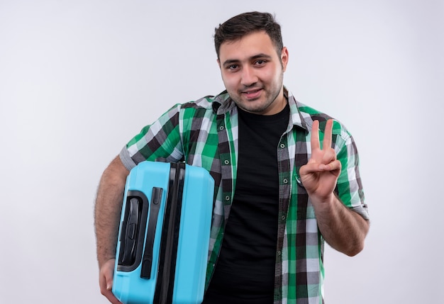 Homme jeune voyageur en chemise à carreaux tenant valise montrant et pointant vers le haut avec le doigt numéro deux souriant debout sur un mur blanc