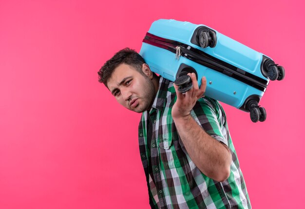 Homme jeune voyageur en chemise à carreaux tenant une valise sur ses épaules à la fatigue et s'ennuie debout sur un mur rose