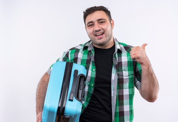 Homme jeune voyageur en chemise à carreaux tenant valise souriant joyeusement pointant vers l'arrière debout sur un mur blanc