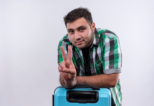 Homme jeune voyageur en chemise à carreaux avec valise montrant le signe de la victoire souriant confiant debout sur mur blanc