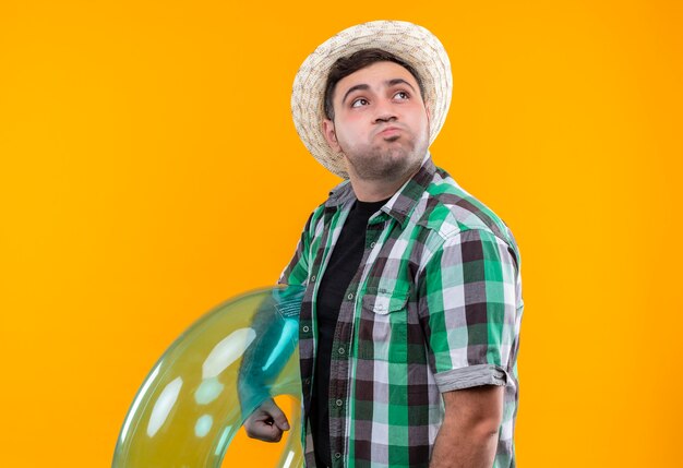 Homme jeune voyageur dérangé en chemise à carreaux et chapeau d'été tenant l'anneau gonflable à côté soufflant les joues debout sur le mur orange