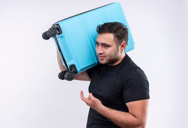 Homme jeune voyageur en t-shirt noir tenant valise à côté confus et très anxieux debout sur un mur blanc